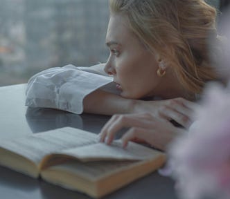 A forlorn woman leans over a book and looks out a window