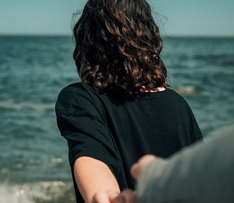 Photo of a woman walking away from the camera, towards to water and blue sky. Her arm is behind her, holding the hand of someone who’s are is reaching forward to her.