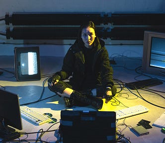 A woman in wearing a black jacket is sitting on the floor connected to various screens and keyboards.