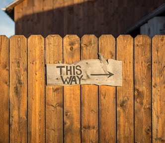 A rustic fence made of brown wood holds a sign that says “THIS WAY” with an arrow pointing to the right.