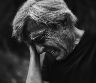 A black and white close-up portrait of an older man bawling out, crying as he cups his hands on his forehead