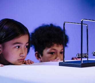 two children stating at a Newton’s cradle or pendulum — a device that typical sits on desktops, consisting of five metal hanging balls. when one ball is pulled sideways then released, it collides with the other balls, which forces the ball on the other side into motion, thus starting a back-and-forth chain reaction that, while not literally “perpetual,” tends to continue for a very long time.