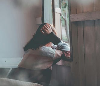 A woman leans against a windowsill in quiet contemplation, resting her head on her hand. Soft light filters through an open wooden window, creating a calm, introspective atmosphere.