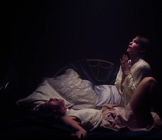 Still from “A Banquet” featuring Betsy played by Jessica Alexander and Holly played Sienna Guillory. Both are in a dark room with a single light overhead. Holly is praying while Betsy is on her back screaming.