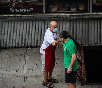A NYC shopkeeper and a customer, both wearing masks, bump elbows in greeting on the sidewalk outside the shop.