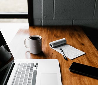 Wooden table with opened MacBook, white coffee mug filled with black coffee, a notepad with black pen and a smartphone on it.