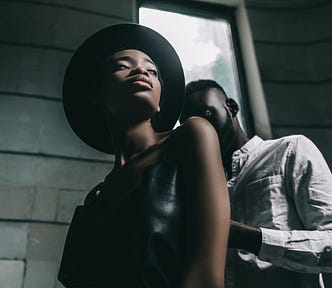 Black man behind Black woman in front of concrete wall and window