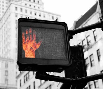 Photo of a “stop” hand lit up on a crosswalk sign. White, city buildings are in the background.