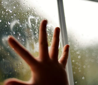 Childs hand pressed against window in home quarantine lockdown with rain drops and green leaves outside.