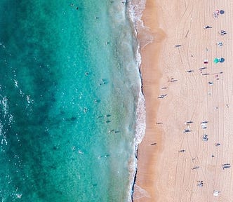 overhead drone shot of beach, turquoise sea at left, sand at right, waves breaking on shore, people dotted along the sand