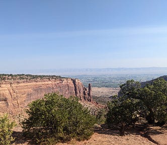 A vista from Colorado National Monument. A giant outcropping of rock takes up the left side, with smaller pieces at the end that break away from the main rock. Mountains can just barely be seen on the far horizon under a beautiful blue sky with wispy clouds. Shrubs dot the foreground.