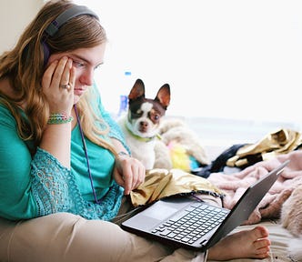 Young, overweight, white woman on bed typing on laptop with small dog looking over shoulder