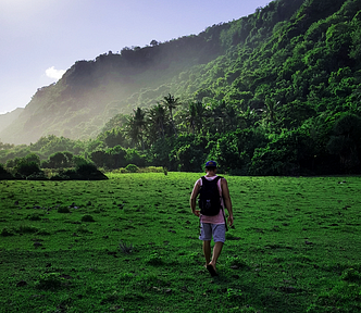 A man wearing a backpack hikes across a field toward a vegetation covered mountain.