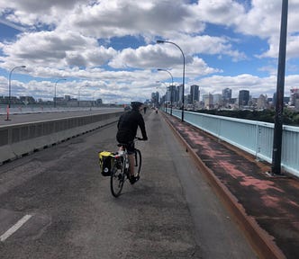 A photo of a young man on a bicycle, riding along a long highway, with a blue sky dotted with clouds above, and a city in the distance ahead