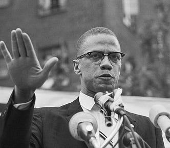 Black and white photograph of head shot of Malcolm X with hand raised in front of microphones