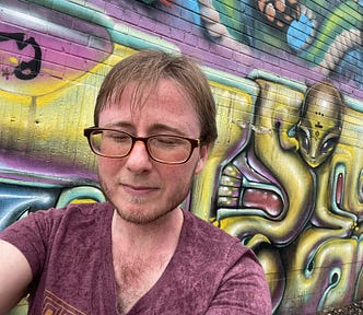 The author is standing in front of a wall covered in colorful graffiti. It’s raining. He has his eyes closed and is not smiling. He has glasses, short brown hair, and a beard. The graffiti includes an alien from outer space. The rest is abstract art.