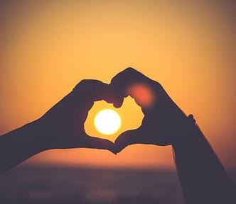Two hands form a heart shape, in a silhouette set agains the setting sun. The photo is a monotone of yellow and orange colors, with a vignette darkening the border.