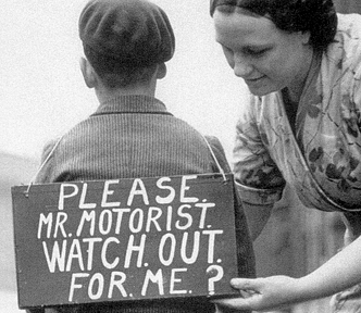 A mother attaches a sign to her son getting ready to ride his bicycle. It reads: ‘Please Mr Motorist. Watch out for me?’