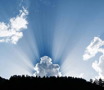 Sun rays radiating upward from behind a puffy cloud behind a treeline.