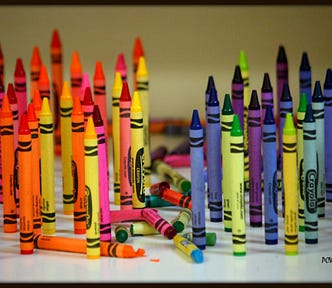 Image is titled Crayon Wars. There are around sixty crayons standing with points upward. The crayons are divided, warm color crayons (red, orange, yellow) on the left and cool color crayons (blue, green purple) on the right. In the middle is a pile of broken crayons, warm and cold. It’s a war, or maybe a waxing.