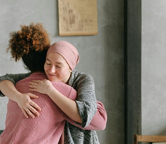 Two women are embracing in a grey walled room. The face of the woman we can see is smiling.