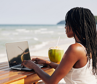 Black woman with long, natural locked hairstyle, typing on a laptop at the beach, drinking from a fresh coconut