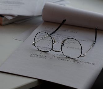 pair of glasses laying on a pad of paper with writing on it — you should proofread your Medium stories.