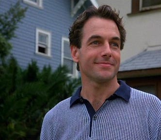 A color image from The Deliberate Stranger, showing Mark Harmon with a smile as he portrays the charming side of Ted Bundy. He has tussled dark hair and blue eyes, and he is wearing a shirt with a dark blue collar and a blue and white pattern. Houses are visible behind him.