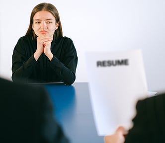 A woman with her elbows on the table and hands under her head, looking pensively at another person holding a piece of paper that says Resume