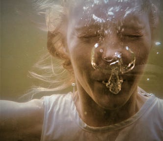 A photograph of a small child under the water with her eyes squeezed shut and surrounded by the air bubbles she is blowing out.