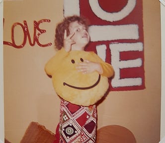 Author at age 3 — standing on her bed holding a yellow smiley face pillow while making the peace sign with her two fingers. She’s wearing a yellow shirt and wildly patterned pants. The wall behind her has a glass sign that says “Love” and also a shag latch hook rug that says “Love”