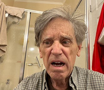 A man with disheveled gray hair and a plaid shirt in a bathroom with a surprised or distressed facial expression. The background includes a glass shower door, a beige towel, and a red garment hanging on a hook. A squeegee can be seen inside the shower.