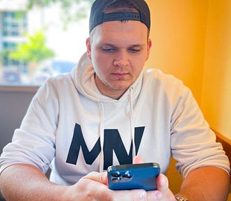 Isaac Mashman wearing a backward hat and white Mashman Ventures hoodie, sitting in front of an orange wall, looking at his phone.