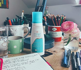 A bottle of Estrogel (oestrogen) on a desk surrounded by a coffee cup, glass of water, a mug of pencils and pink flowers.
