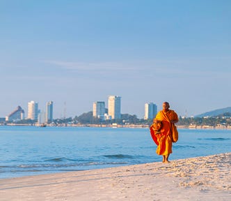 An orange-clad monk walking toward the camera on the beach, with sparse buildings on the horizon under a vast blue sky.