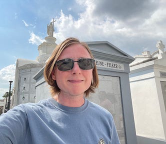 The author is standing in front of some graves in the center of New Orleans. He is wearing sunglasses and a light blue t-shirt.