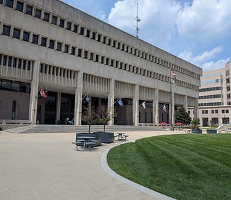 The front of the Towson Courthouse (Baltimore County Courts Building). A large light tan walkway is visible leading up to the steps, with a circular expanse of well-kept grass just to the front right. The building is a large modern building, light tan with columns and flags in the front.