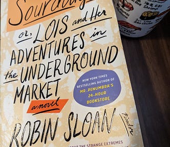 A copy of Sourdough or Lois and Her Adventures in the Underground Market by Robin Sloane sits on a table with a Japanese-style tea mug (tea visible inside) with depictions of various kinds of sushi to the right of the book.