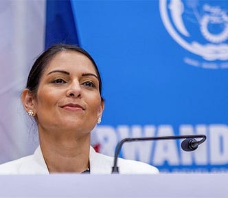 Preti Patel looking smug in front of the microphone and a poster with Rwanda written on it.