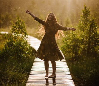 A woman dances in a spring rain-shower with a smile on her face and her arms spread wide. The sun shines through the rain.