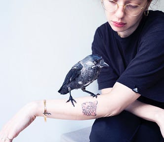 A woman wearing glasses and a tiger tattoo, with a crow standing on her arm.