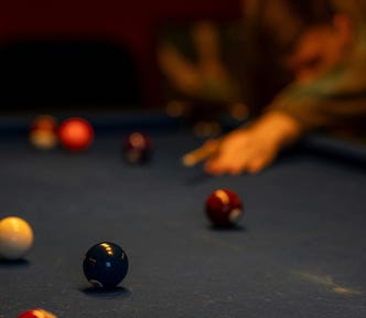 A man leans over a dimly lit pool table, cue stick poised to strike. The scattered balls mirror a life out of order — precision and chaos suspended in a single breath before the break.