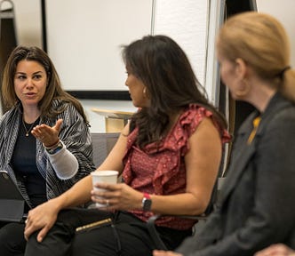 One woman on the left is gesturing while speaking, and the other two women are listening attentively. They all hold coffee cups, and the setting appears informal yet professional, with a flip chart visible in the background.