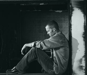 A black and white photo of a man in an old, worn down basement looking extremely depressed.