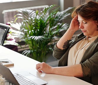 A middle age woman sitting at a desk with a laptop with her right hand holding her head in apparent discomfort