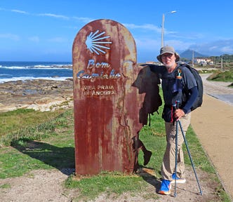 Man wearing a cap, backpack and carrying hiking poles leans against a rusted metal sign with a cutout of the Camino scallop shell and the words Bom Cominho and Vila Praia de Ancora. (Good Journey and Village Beach of Ancora)