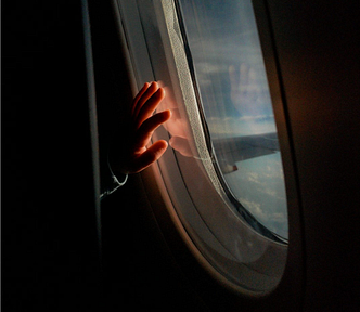A small child’s hand pressed against an airplane window, backlit by cloudy sky, the foreground ominously dark