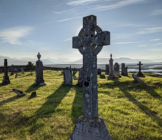 Crosses and tombstones with mountains beyond and low light create shadows on the grass of the cemetery in a wild Scotland landscape