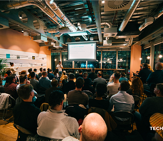 Chuck speaking with a microphone, presenting Design Sprints on a projector to a live audience at WeWork Spinningfields, Manchester. Room is softly lit, and has an industrial style.