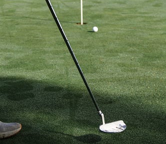Ball approaches a hole with pin in, putter head and shaft in foreground